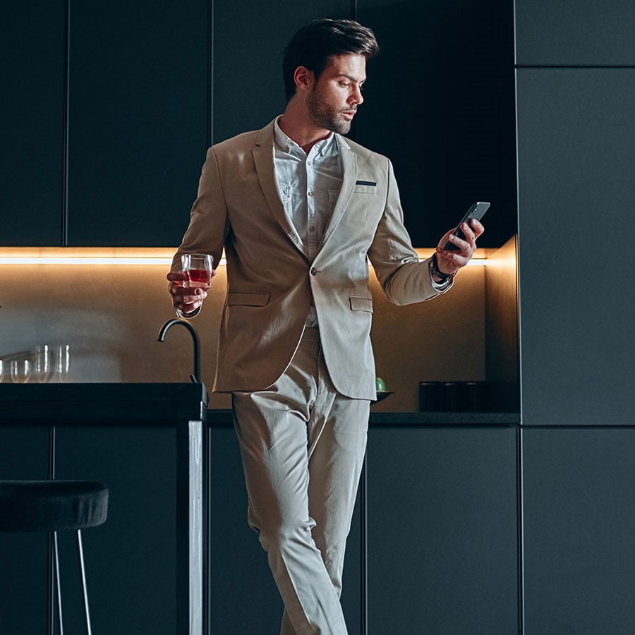 A man in a beige suit leans against a sleek, modern kitchen counter, holding a smartphone and a glass of wine, with a relaxed, sophisticated vibe.